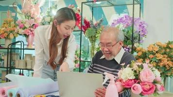Asian elder male florist owner discussing with young beautiful female employee who shopkeeper about website arrangement for business service, happy work in colorful flower shop store, e-commerce SME.