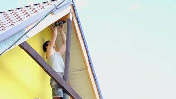 Male builder makes exterior decoration of private house in the style of half-timbered house with his own hands - he mounts wooden beams on facade, pediment under the roof high on the scaffolding