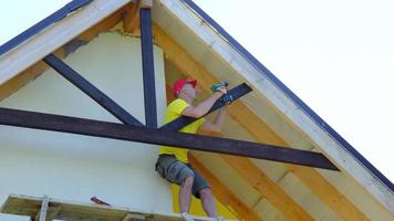 Male builder makes exterior decoration of private house in the style of half-timbered house with his own hands - he mounts wooden beams on facade, pediment under the roof high on the scaffolding video