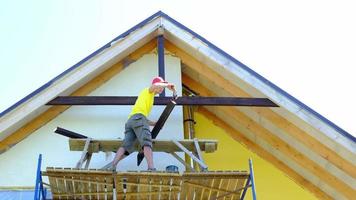 Male builder makes exterior decoration of private house in the style of half-timbered house with his own hands - he mounts wooden beams on facade, pediment under the roof high on the scaffolding video