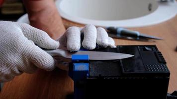 verscherping een mes Aan een elektrisch slijper Bij huis. de man's hand- schijven de mes blad tussen de blauw slijpers, stof vliegt Aan de machine. 4k, langzaam beweging video