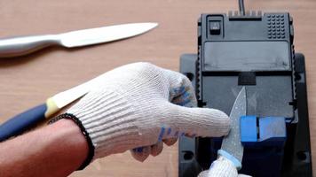 Schärfung ein Messer auf ein elektrisch Anspitzer beim heim. das des Mannes Hand fährt das Messer Klinge zwischen das Blau Spitzer, Staub fliegt auf das Maschine. 4k, schleppend Bewegung video