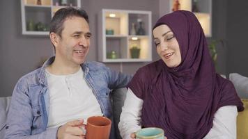 Happy Muslim couple having intimate conversation at their home. The headscarved woman and her husband are sitting on the sofa at home, drinking coffee and chatting.