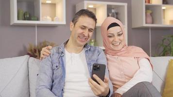 musulmán familia reír, mirando a el teléfono y teniendo divertida. musulmán mujer en hijab y su marido sentado en sofá a hogar y mirando a teléfono y reír. video