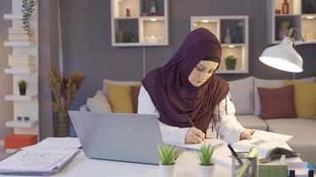 musulmán mujer en hijab trabajos a hogar oficina. musulmán mujer trabajando en hogar oficina trabajos desde ordenador portátil y toma notas video