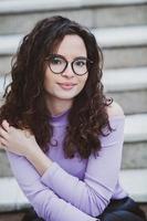 Beautiful young woman with brunette curly hair, portrait in eye glasses enjoying the sun in the city. photo