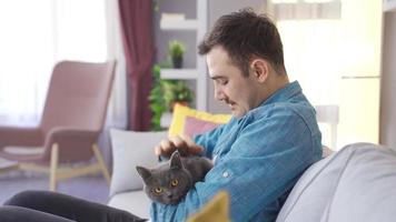 The man loves his cat at home and strokes the cat's head. The man sitting on the sofa with his gray cat loves the cat and strokes it with his hand. video