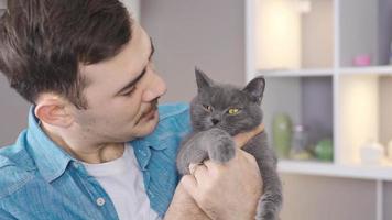 The man holds his gray cat in his lap and looks at him affectionately. Close-up of man holding a gray cat in his arms with a strong hug.