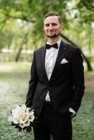 groom in a black suit with a bow tie in a city park photo