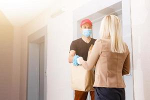 Young woman wearing medical mask receiving orderd food from the restaurant from delivery man indoors. Prevention of virus spread photo