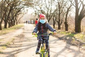 pequeño niña vistiendo médico máscara evitar gripe, contaminaciones y covid-19 montando bicicleta exterior. foto
