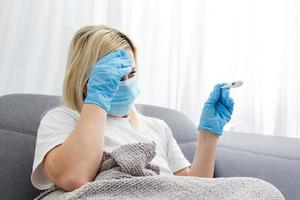 Woman in medical mask measures body temperature. Sick girl looks at digital thermometer in her hands, concept of cold and flu, fever and coronavirus symptoms photo