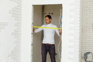 Master builder in uniform of measuring distances on a white wall with a tape measure photo