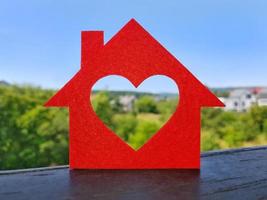 Small house cut out of red felt is standing on balcony railing. Heart-shaped hole is cut instead of a window. Blurred background of cottage village in summer sunny day. Dream home concept. photo