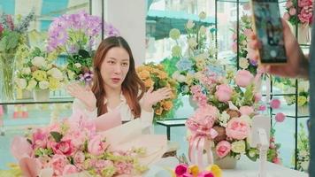 e-handel företag, ung asiatisk kvinna blomsterhandlare arbetstagare demonstrerar och visar blommig arrangemang via uppkopplad leva strömning förbi smartphone Ansökan i ljus blomma affär, skön blommar Lagra. video