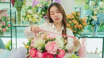 One Young beautiful Asian female florist entrepreneur arranging a bunch of blossoms, decorating with lovely ribbons, happy work in colorful flower shop store with blooms, and small business owner. video