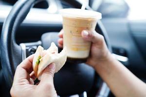 Asian woman driver hold ice coffee cup and sandwich bread for eat and drink in car, dangerous and risk an accident. photo