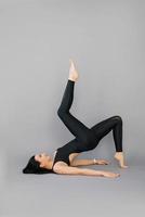 Young woman fitness trainer or yoga performs an exercise lying on her shoulder blades and lifting one leg at the top on a gray background photo