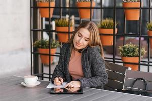 Young charming woman is calling on a mobile phone, sitting in a cafe and talking on a mobile phone with a smile, writing notes in a notebook. Remote work photo