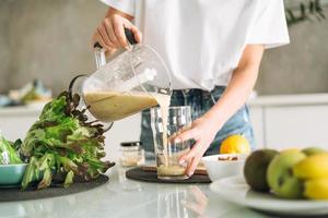 Young slim woman in white t-shirt and blue jeans cooking green smoothie healthy food in kitchen at home photo