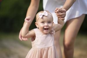 A happy one-year-old girl is holding her mother's hands. Little child for a walk. Sweet baby learns to walk. photo