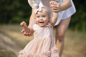 A happy one-year-old girl is holding her mother's hands. Little child for a walk. Sweet baby learns to walk. photo