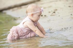 A beautiful little girl sits on the river bank and plays with sand and water. Child and nature. photo