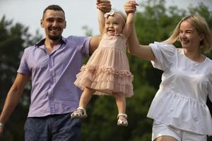marido, esposa y su pequeño hija para un caminar en el parque. contento papá y mamá lanza arriba su pequeño riendo un año de edad hija. foto