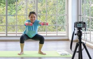 Happy young asian woman lifting dumbbells exercise and live streaming with smiling face. Beautiful smile young Asian woman exercise at home. Young people domestic life concept. Selective focus. photo