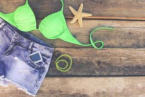 Summer women's clothing and accessories bathing suit, denim shorts, mobile phone, headphones, shells. Toned image. Top view. photo