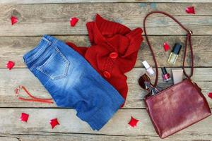 Women's autumn clothing and accessories red sweater, jeans, handbag, beads, sunglasses and cosmetics on wooden background. Top view. photo