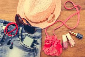Summer women's accessories sunglasses, beads, denim shorts, mobile phone, headphones, a sun hat, handbag, lipstick, nail polish. Toned image. Top view. photo
