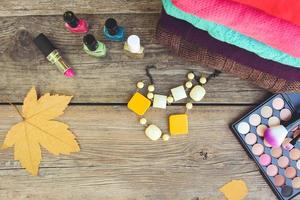 Women's clothing and cosmetics sweaters, lipstick, nail polish, necklaces, eye shadow, brushes, yellow leaves on wooden background. Top view. Toned image. photo