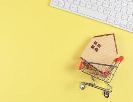 flat layout of wooden house model in shopping trolley and  and computer keyboard  on yellow background with copy space, home purchase concept. photo