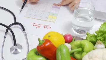 nutricionista mulher prepara dieta plano para saudável vida às mesa. nutricionista mulher escrevendo dieta plano em mesa cheio do frutas e legumes. video