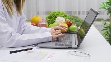 trabajando con computadora portátil, dietético prepara investiga y planes para sano viviendo. nutricionista mujer mecanografía dieta plan en ordenador portátil a mesa lleno de frutas y vegetales. video