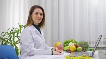 positivo dietético mujer trabajando a mesa lleno de vegetales y frutas sonriente a cámara. dietético mujer satisfecho con su trabajo mirando a cámara y sonriente. video