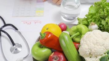 nutritionniste femme l'écriture régime plan sur table plein de des fruits et des légumes. femelle nutritionniste en utilisant portable dans Bureau prépare régime planifier. video