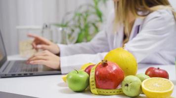 dietologo donna Lavorando con il computer portatile nel Ospedale. frutta e verdure su il tavolo. dietologo donna Lavorando su il computer portatile nel ospedale e preparazione salutare mangiare piani. video