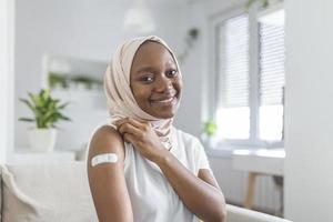 retrato de un musulmán africano hembra sonriente después consiguiendo un vacuna. mujer participación abajo su camisa manga y demostración su brazo con vendaje después recepción vacunación. foto