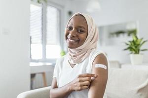 retrato de un musulmán africano hembra sonriente después consiguiendo un vacuna. mujer participación abajo su camisa manga y demostración su brazo con vendaje después recepción vacunación. foto