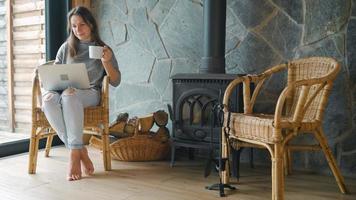 mujer utilizando ordenador portátil y Bebiendo café sentado cerca el chimenea. video