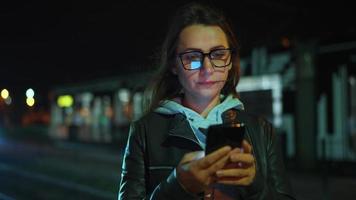 vrouw staat Bij een vervoer hou op Bij nacht, gebruik makend van smartphone en aan het wachten voor tram video