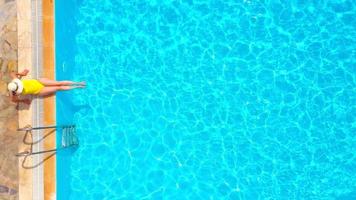 Top view of a woman in yellow swimsuit and a hat sits on the edge of the pool video