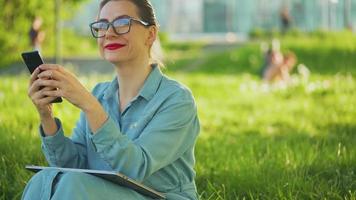 mujer utilizando teléfono inteligente mientras sentado en parque después refinamiento al aire libre trabajo video