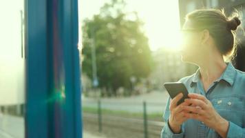 Woman stands at a transport stop, using smartphone and waiting for the tram. video