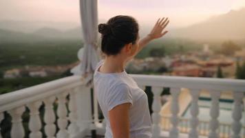 mujer va fuera en el terraza mirando fuera terminado puesta de sol. video