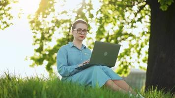 Busy attractive woman working at the laptop as sitting on grass in city park video