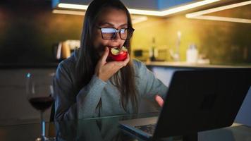 Woman is sitting in the kitchen and working on a laptop and eating the apple video