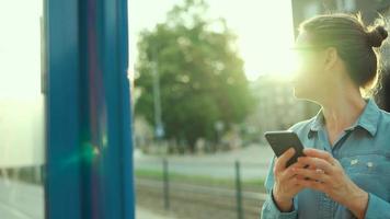 vrouw staat Bij een vervoer hou op, gebruik makend van smartphone en aan het wachten voor de tram. video